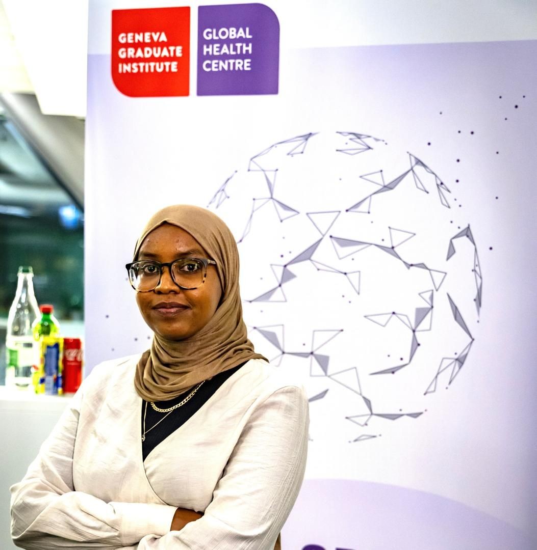 Person in a hijab standing confidently in front of a Geneva Graduate Institute banner indoors.