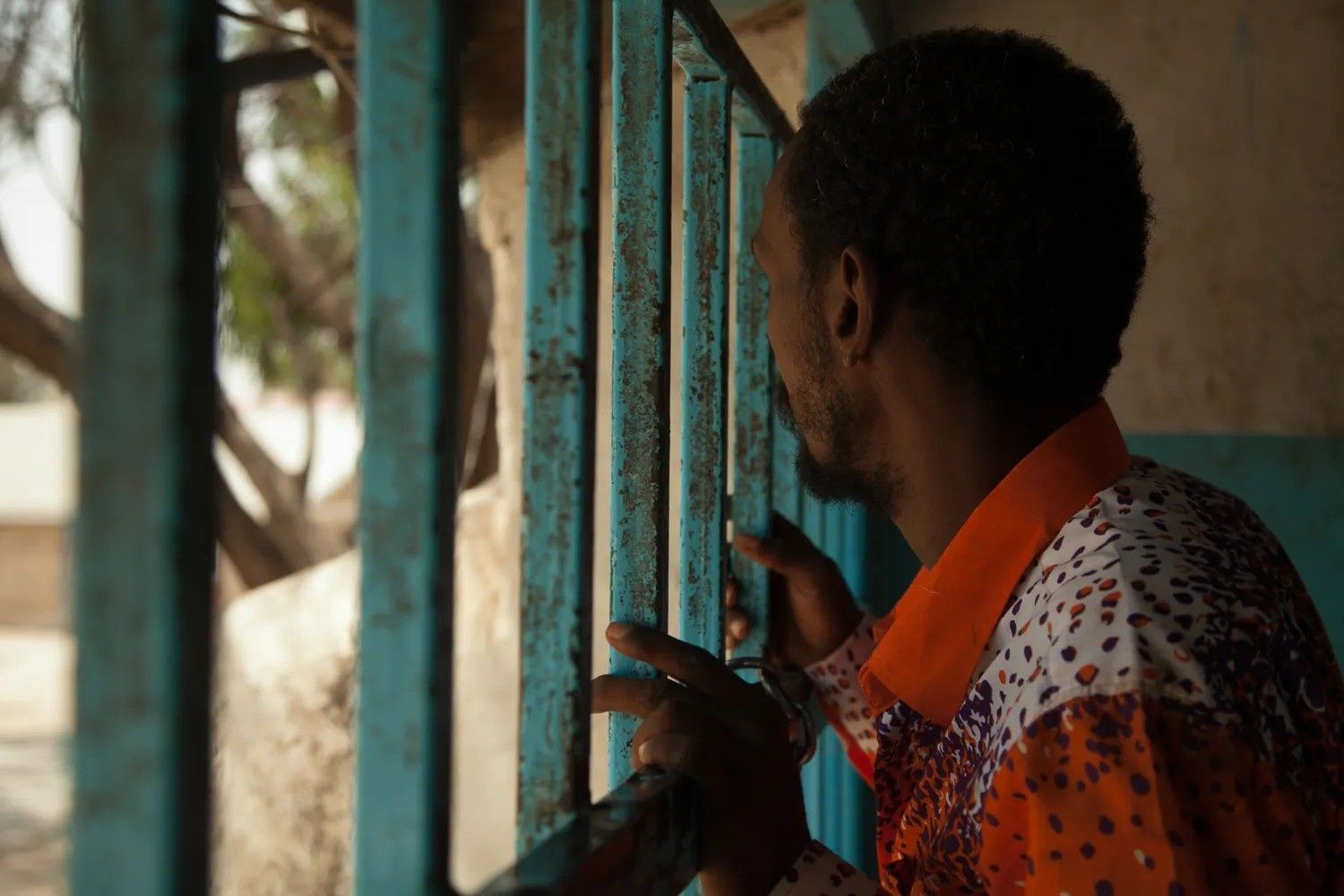 Person in a colorful shirt looking through blue rusted bars, with hands gripping the bars.