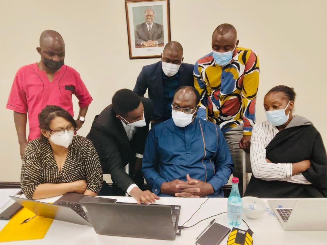 Group of people wearing masks gathered around a table with laptops, having a discussion in an office.