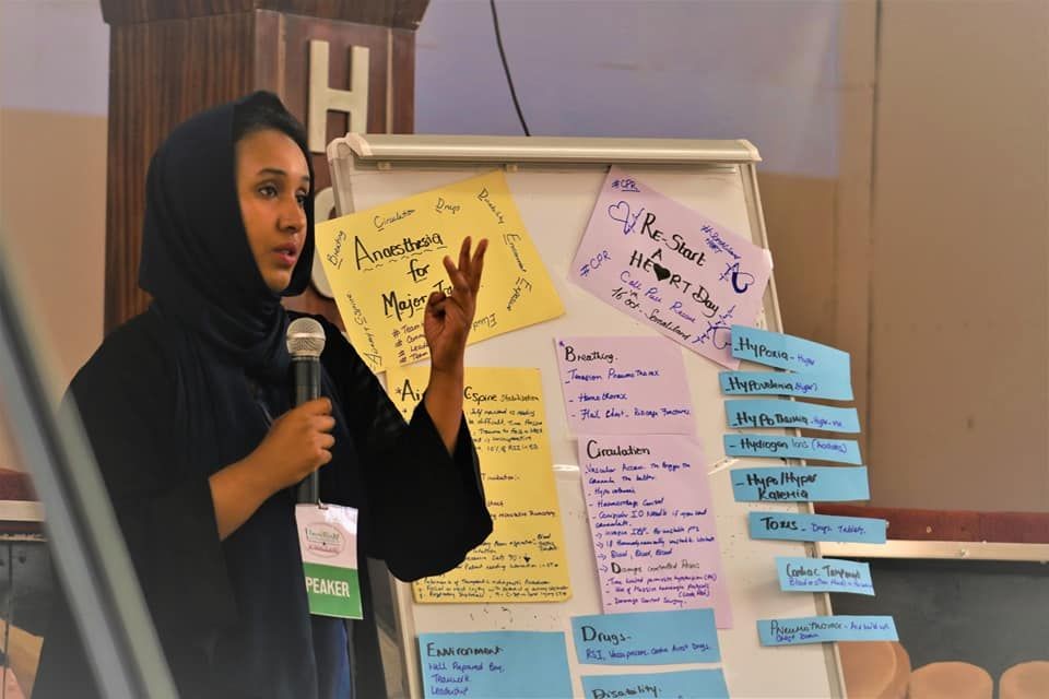 Speaker giving a presentation with notes about anesthesia and medical topics displayed on a flipchart.