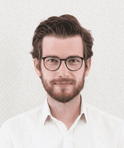 Man with glasses and a beard wearing a white shirt, standing against a patterned background.