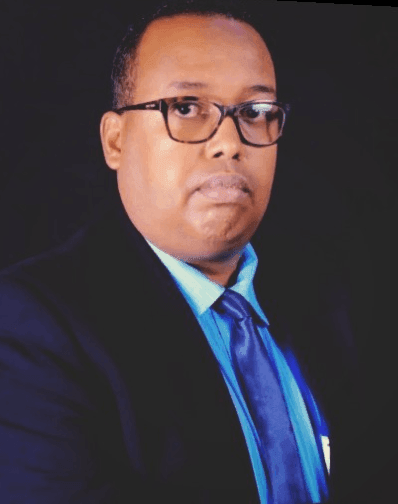 Person in a suit with a blue shirt and matching blue tie, against a dark background.