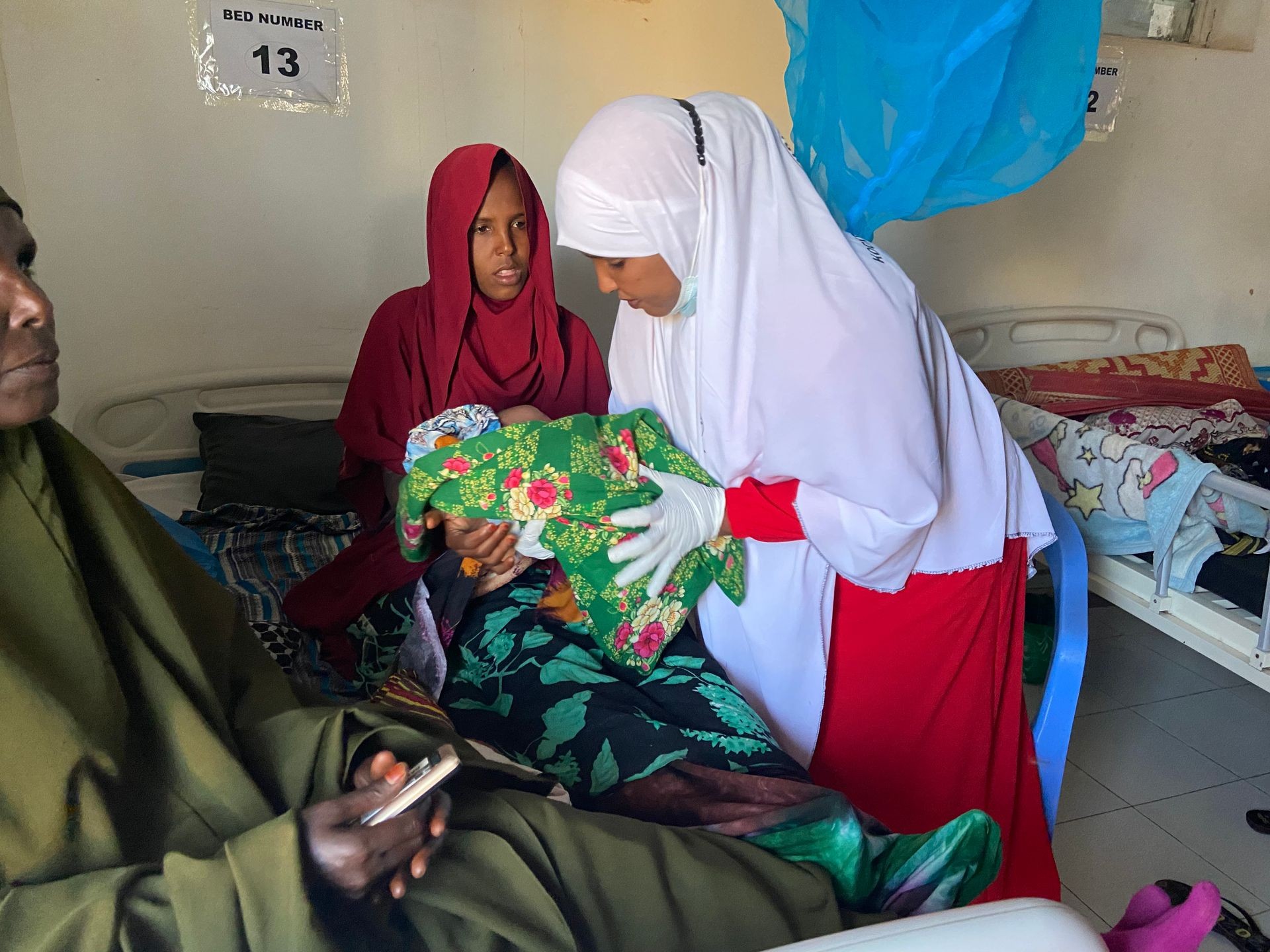 A healthcare worker in white and red attire assisting a mother holding her baby in a hospital room.