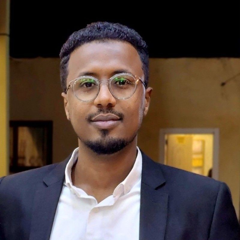 Person wearing a white shirt and black blazer standing indoors with a room in the background.